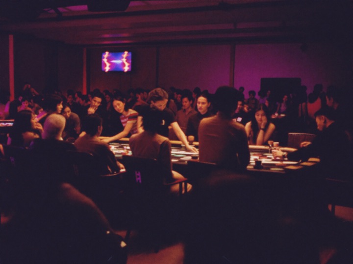 A busy live casino room at night, guests standing around gaming tables under low lighting