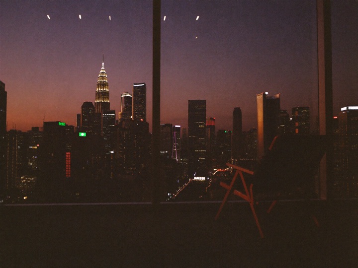 A Kuala Lumpur skyline at dusk seen from a high window, an empty lounge chair in front