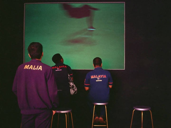 A sports lounge wall of large screens, three guests watching from bar stools